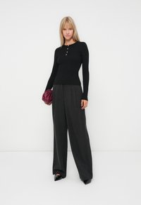 Zwarte ribbel top met lange mouwen en knoopdetails, gecombineerd met wijde charcoal grijze broeken en een gekreukelde bordeauxrode clutch. Zwarte puntige schoenen.