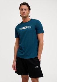 Turbogruen bomullströja med rund halsringning och korta ärmar; har en iögonfallande vit "HUMMEL"-logotyp över hela framsidan; bärs med svarta shorts.