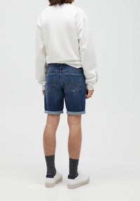 Shorts en denim de couleur foncée avec des revers roulés. Ils comportent deux poches arrière avec des coutures discrètes. Portés avec un sweat-shirt blanc et des baskets blanches.