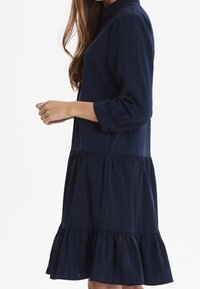 Abito blu navy con orlo a strati e ruffle, maniche lunghe e polsini abbottonati. Realizzato in tessuto leggero con una texture morbida.