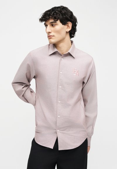 Νέος άνδρας με σγουρά σκούρα μαλλιά, ντυμένος με ροζ ανοιχτό πουκάμισο με κουμπιά και λογότυπο "EA" στο στήθος, κοιτάζοντας πλάγια.