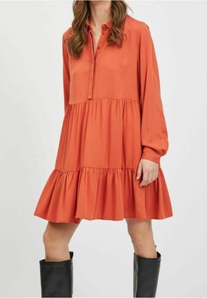 Femme portant une robe orange ample à manches longues et à volants avec un col, associée à des bottes noires montantes jusqu'aux genoux.