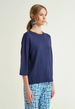 Femme aux cheveux blonds portant un haut bleu foncé à manches trois-quarts, de grands boucles d'oreilles bleues, et un pantalon à motif bleu, debout devant un fond clair.