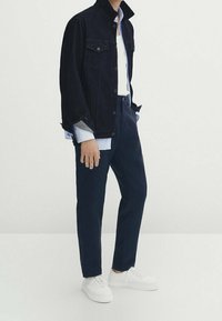 Mörk denimjacka med två bröstfickor, vit t-shirt och randig skjorta under. Mörkblåbyxor och vita sneakers fulländar outfiten.