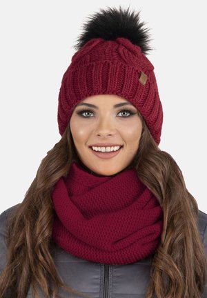 Berretto a maglia bordeaux con pompon in pelliccia sintetica nera sulla sommità. Accompagnato da una sciarpa a trama coordinata dello stesso colore.