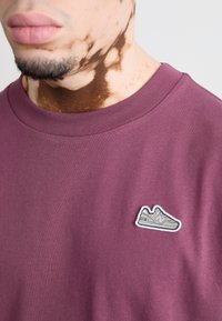 T-shirt di cotone color bordeaux con colletto a girocollo a costine. Presenta una piccola patch a forma di sneaker in beige e grigio sulla parte sinistra del petto.