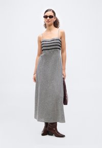 Γυναίκα που φοράει μακρύ φόρεμα με ασπρόμαυρο καρό μοτίβο και λεπτές τιράντες, σκοτεινά γυαλιά ηλίου, καφέ μπότες και κρατά μια καφέ τσάντα.