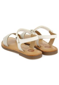 Sandalias de piel blanca con correas trenzadas, suela plana marrón y correa de tobillo ajustable. Presenta un forro interior suave y un detalle de logo.