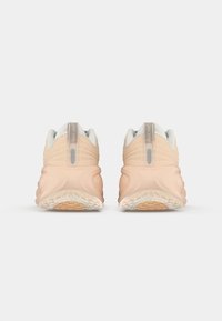 Pantofi sport de culoare piersică deschisă, cu partea superioară din plasă texturată, guler căptușit, talpă groasă și sistem de dantelare vizibil; includ o etichetă pe călcâi semi-transparentă.