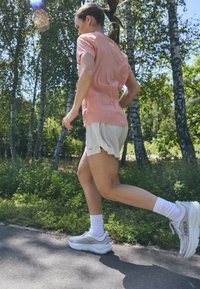Svijetlopinkasta majica s kratkim rukavima, bijeli sportski šortsevi i sivi tenisice s amortizacijom, postavljeni protiv zelene, šumske pozadine.