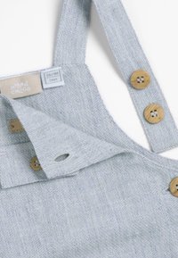 Salopette pour bébé en tissu bleu clair texturé avec des boutons en bois et une étiquette indiquant la taille 56/1M pour un bébé d'1 mois.