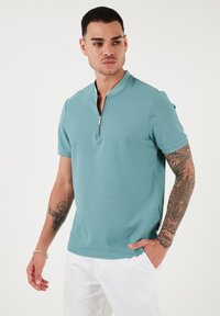 Turkizna polo majica s kratkimi rokavi in pol-zadrgo; teksturirana tkanina s prilegajočim oblikovanjem in gladkim zaključkom. Spodaj nosite bele hlače.
