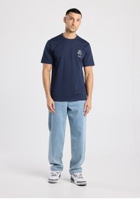 T-shirt en coton bleu marine à manches courtes avec un motif d'arbre sur le côté gauche de la poitrine, accompagné d'un jean en denim bleu clair et de baskets blanches.