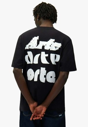 Homme à la peau foncée avec de courtes dreadlocks porte un t-shirt noir avec un grand texte blanc déformé dans le dos, les mains jointes derrière.