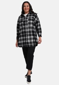 Veste à carreaux noir et blanc avec une coupe décontractée, boutons à l'avant et col. Portée sur un pantalon noir et des sneakers noirs.