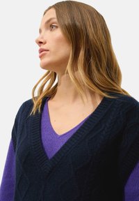 Granatowy sweter w warkoczowy splot, nałożony na fioletową bluzkę z dekoltem w serek, z prążkowanym wykończeniem i strukturalnym wzorem dzianiny.