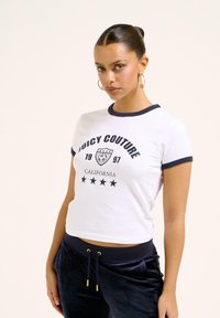Balta trumpa marškinėlių su mėlynomis dekoracijomis, ant kurių yra "Juicy Couture" tekstas, skydo grafika ir penkios žvaigždės po ja.