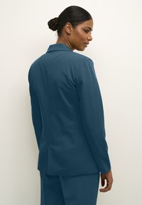 Tyrkysový blazer na míru se skrytým rozparkem, slim fit, a strukturovanými rameny. Hladká tkanina s jemným leskem, doplněná o kalhoty ve stejném odstínu.