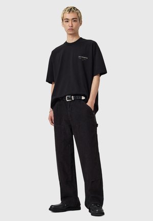 T-shirt noir surdimensionné avec logo sur le devant, associé à un pantalon en velours côtelé noir avec poches et ceinture. Les chaussures sont noires avec des bouts arrondis.