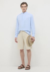 Camicia azzurro chiaro a maniche lunghe con colletto alla coreana e apertura con bottoni, pantaloni beige, sandali marroni e cappello di paglia con un nastro a righe.