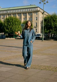 Femme portant une veste en jean et un jean marchant en plein air tenant un plateau avec une tasse de café et un croissant dans une place urbaine par une journée ensoleillée.