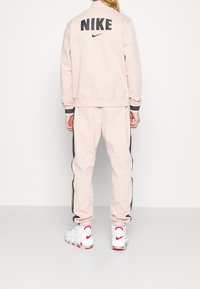 Rosa Nike träningsoverall med rund halsringning, svart logotyp på ryggen och kontrasterande sidostråkar. Matchad med vita sneakers med röda detaljer.