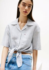 Camisa de botones en azul claro, con rayas blancas, un bolsillo en el pecho y un dobladillo anudado. Combinada con pantalones cortos de mezclilla de cintura alta.
