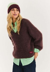 Nadrozměrný, tmavě vínový svetr s načechranou strukturou, vrstvený přes světle zelenou košili. Svetr má malý štítek na lemu jako detail.