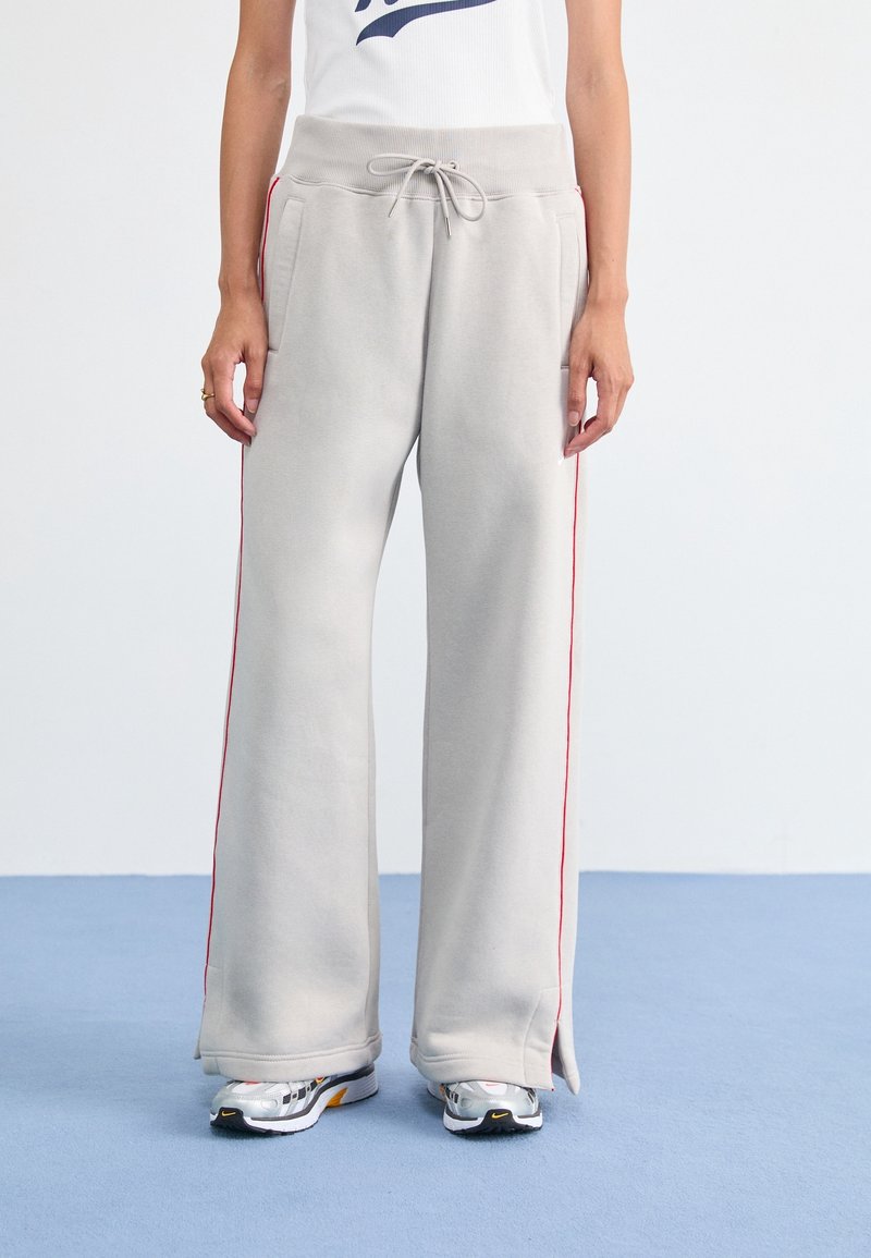 Person som bär beigefärgade vida sweatpants med röda sidostråk och vita sneakers, stående på en blå golv mot en vit vägg.