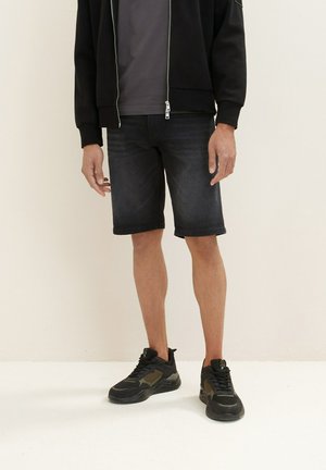 Veste zippée noire, t-shirt gris, shorts en denim foncé et baskets noires avec des accents olive. Coupe droite et longueur courte.