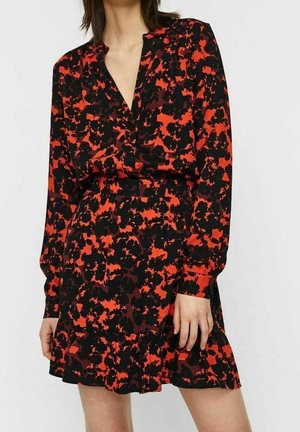 Kobieta w długim rękawie, krótkiej sukience z czerwono-czarnym abstrakcyjnym kwiatowym wzorem i kołnierzykiem, stojąca na jednolitym tle.