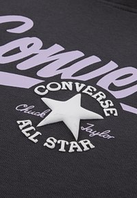 Μαύρο φούτερ από ύφασμα με μεγάλο μωβ κείμενο "Converse" και λευκό λογότυπο αστέρι με το κείμενο "Chuck Taylor All Star" από κάτω.