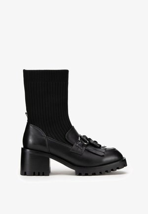 Černé kotníkové boty s ribbovaným sock-like svrškem, koženým tělem, dekorativními třásněmi a robustním gumovým podpatkem s texturovanou podrážkou.