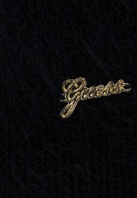 Tessuto nero felpato con una spilla in oro con il logo "Guess", caratterizzato da un font corsivo e un accento a forma di cuore. La consistenza è morbida e soffice.