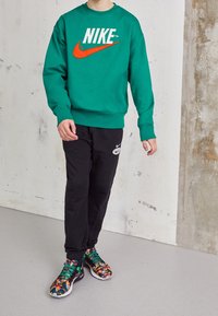Person med grön Nike-tröja, svarta Nike-joggingbyxor och färgglada mönstrade sneakers står mot en texturerad vägg.