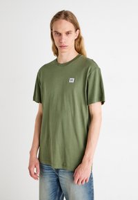 T-shirt in cotone verde oliva con scollo rotondo, maniche corte e una piccola etichetta cucita sul petto. Vestibilità casual, texture morbida.