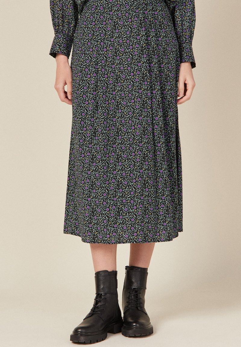 Schwarzer Midirock mit floralem Muster, der ein lila und grünes botanisches Design zeigt. Kombiniert mit schwarzen Schnürkampfstiefeln. Weicher, fließender Stoff.