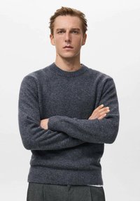 Giovane uomo con capelli castano chiaro che indossa un maglione di lana grigio scuro, in piedi con le braccia incrociate su uno sfondo bianco semplice.