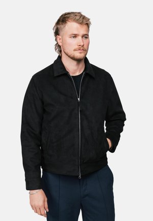 Jeune homme aux cheveux châtain clair portant une veste noire zippée et un pantalon bleu foncé, debout avec une main dans la poche sur fond blanc.