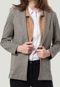 Szarek marynarka w kratę z wzorem houndstooth w kolorze czarno-białym, z tanowym futrzanym podszyciem i klapami, noszona na białej koszuli.