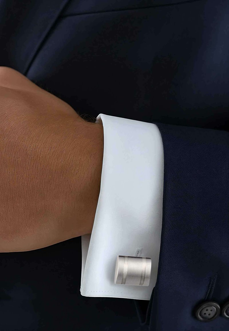 White cuff of a dress shirt with a silver cylindrical cufflink on a dark suit sleeve, showcasing smooth fabric and a clean design.