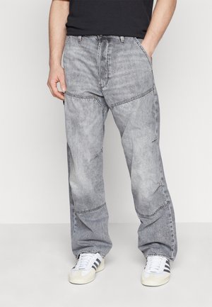 Mężczyzna w szarych, spranych luźnych dżinsach i białych butach sportowych z czarnymi paskami, stojący na tle jasnego, jednolitego tła.