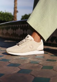 Beige pletené tenisky s texturovaným svrškem, bílou podrážkou a zlatými akcenty. Mají logo na boku a zaoblený design špičky.