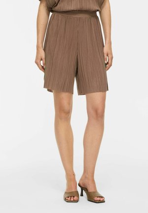 Femme portant un short plissé marron mi-genou et des mules taupe à bout ouvert sur un fond uni.