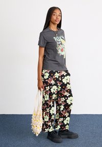 Tricou gri cu un grafic floral, asortat cu pantaloni negri cu modele florale galbene și maronii. Ținând o geantă împletită crem, purtând pantofi negri.