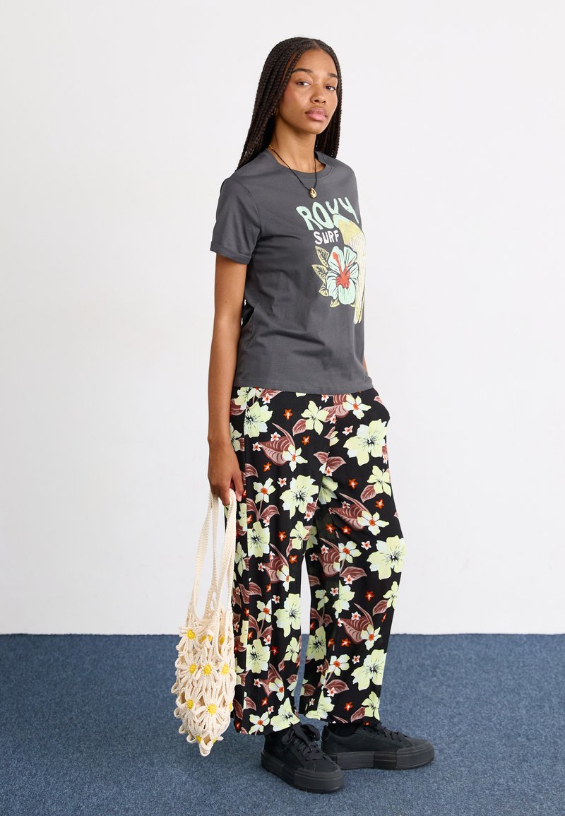 Graues T-Shirt mit floralem Grafikdesign, kombiniert mit schwarzen Hosen mit gelben und braunen Blumenmustern. Hält eine beigefarbene geflochtene Tasche und trägt schwarze Schuhe.