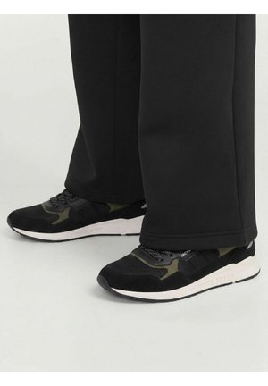 Personne portant des baskets noires et vert olive avec des semelles blanches et un pantalon noir couvrant les chevilles, debout sur une surface claire.