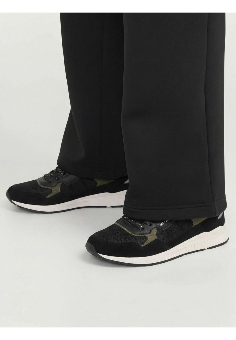 Personne portant des baskets noires et vert olive avec des semelles blanches et un pantalon noir couvrant les chevilles, debout sur une surface claire.