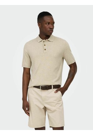 Giovane uomo in piedi con una mano in tasca, indossa una polo beige a maniche corte, pantaloncini kaki chiaro e cintura marrone scuro.
