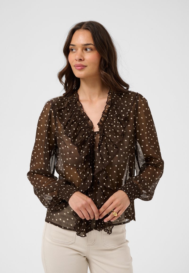 Femme portant un chemisier marron transparent à pois blancs avec un bord volanté, associé à un pantalon clair, debout devant un fond uni.